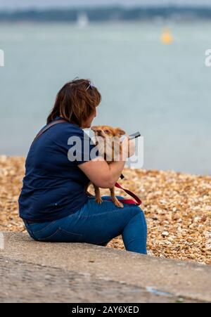 Eine Frau oder Dame, die am Strand am Meer sitzt und einen kleinen Hund hat, der einen Terrier an der Schindelküste hält. Stockfoto