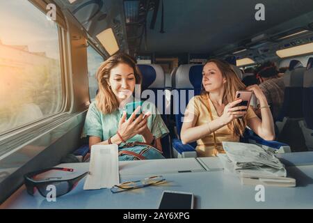 Glücklich sind zwei weibliche Freunde, die mit ihrem Smartphone mit einem langen, weit entfernten Hochgeschwindigkeitszug unterwegs sind Stockfoto