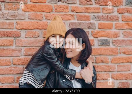 Lächelnde brünette Frau und ihre kleine Tochter umarmen sich mit Liebe, haben ein charmantes Lächeln, tragen schwarze Lederjacken, posieren gegen die Ziegelwand. Junge br Stockfoto