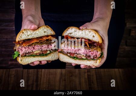 Hand, die einen gespritzter Craft-Rinderburger mit Käse, Speck, karamellisierter Zwiebel und Raketenblättern hält Stockfoto