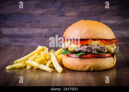 Handwerklicher Rinderburger mit Käse, italienischen Peperoni, Tomaten, Basilikumblättern und pommes frites auf Holztisch und rustikalem Hintergrund Stockfoto