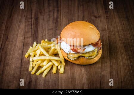 Handwerklicher Rinderburger mit Käse, Ei, Speck, Pickeln und pommes frites auf Holztisch und rustikalem Hintergrund Stockfoto