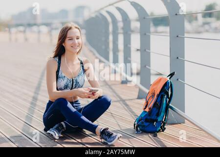 Foto des entspannten, fröhlichen Fitnessmädchens in Sportswear, sitzt im Freien, ruht nach langem gehen, hört Musik mit Handys und Kopfhörern, Ruck Stockfoto