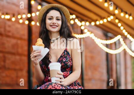 Junges weibliches Modell mit dunklen Augen, rot bemalten Lippen mit Sommerkleid und Hut in den Händen, Kaffee und Croissant zum Mitnehmen, Frühstück im ou Stockfoto