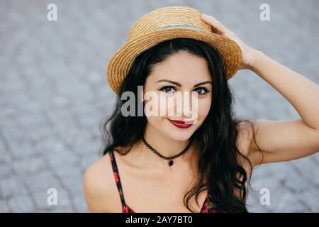 Outdoor-Portrait von brünettem Weibchen mit dunklen Augen, roten Lippen und gesunder Haut mit Sommerhut, wobei sie die Hand am Kopf hält und sich beim gehen ausruhen kann Stockfoto