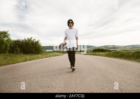 Ein Kerl mit einem Langbrett in den Händen in einem Helm und einer Sonnenbrille läuft auf einer asphaltierten Vorortstraße Stockfoto