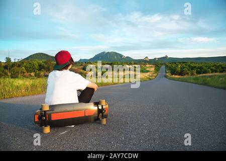 Ein Mann in einer Kappe und mit einem langen Brett in seinen Händen sitzt auf einer Landstraße Stockfoto