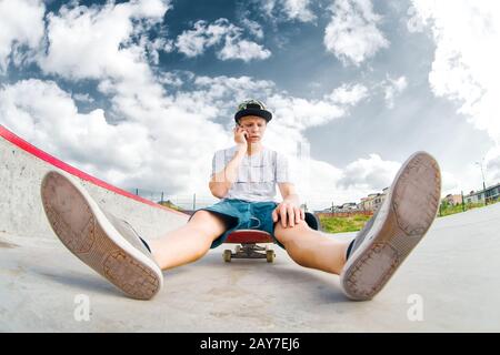Junger Skater sitzt auf Skateboard im Park und telefoniert Stockfoto