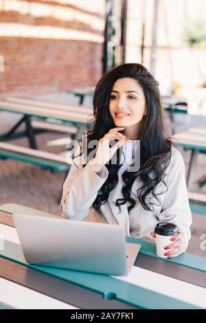 Elegante, erfolgreiche Freiberuflerin, die formell mit einem generischen Laptop für die Arbeit an entfernten Standorten gekleidet war und sich im Café im Freien ausruhen konnte, wo sie köstlichen Kof trinkt Stockfoto