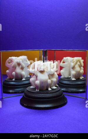 Drei kluge, weiß geschnitzte Affen setzen sich vor einen Doppelspiegel, um alle Posen der Affen zu reflektieren. Blauer Hintergrund, orange und gelb in Spiegeln Stockfoto