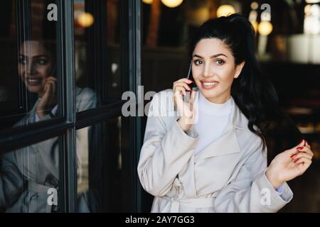 Fröhliche liebenswerte brünette Frau mit ansprechendem Auftritt in weißer Kleidung, die mit ihrem Freund über das Handy klatscht. Junge Geschäftsfrau Stockfoto