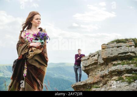 Porträt einer jungen Frau in einem bunten Kleid mit einem Blumenstrauß aus Wildblumen in der Natur, das auf dem Hintergrund ihres Boyfs steht Stockfoto