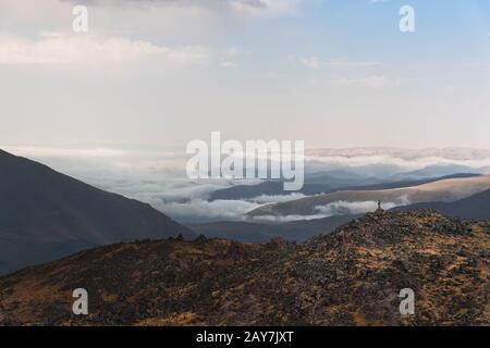 Wanderer der Sieger steht am Rande eines Felsens mit Stöcken, die vor einem von Wolken überzogenen Tal im Kaukasus erhoben werden Stockfoto