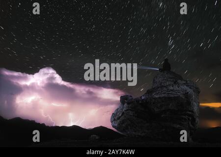 Nachtfoto EIN Mann sitzt auf einem Felsen und strahlt eine Taschenlampe am Himmel, auf der in den Wolken am Sternenhimmel Blitze aufblitzen Stockfoto