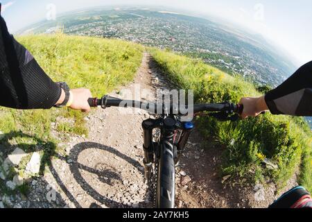 Blick von der ersten Person eines Radfahrers, der von einem hohen Berg im Hintergrund einer Stadt in der Ferne bergab fährt Stockfoto