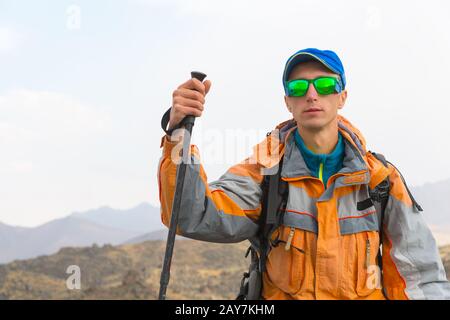 Ein Typ mit einer Sonnenbrille in einer Membranjacke, Kappe, mit Rucksack und Stöcken für Nordic Walking, ein Reisender, der an steht Stockfoto