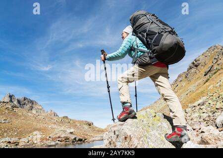 Ein Wandermädchen in Sonnenbrille mit Rucksack und Tracking-Stöcken steigt vor dem Hintergrund von Felsen und einer Höhe von auf einen hohen Felsen Stockfoto