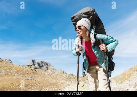 Ein Wandermädchen in Sonnenbrille mit Rucksack und Tracking-Stöcken steigt vor dem Hintergrund von Felsen und einer Höhe von auf einen hohen Felsen Stockfoto