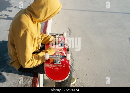 Nahaufnahme eines Teenagers, der in einer Sweatshirt-Jeans und Turnschuhen in einem Skatepark sitzt, der ein Telefon und ein Skateboard hält Stockfoto