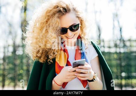 Schöne Frau mit lockigen Haaren, Sonnenbrille und grüner Jacke, die ihr Smartphone in den Händen hält, im Internet surfen und Online-Kommunikation genießen w Stockfoto