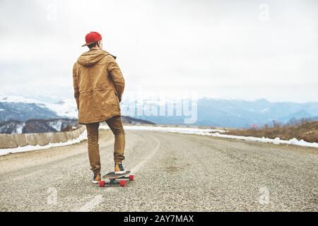 Stilvoller, junger Kerl mit Kappe und Hose Jogger, die auf einem Longboard eine Bergstraße hinunterrollen und so das Leben genießen Stockfoto