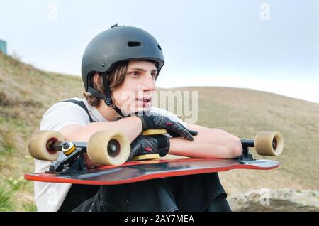 Ein junger Mann in einem Helm, der Handschuhe mit einem Brett in den Händen trägt und gekleidet ist, sitzt auf einem Abgrund hoch in den Bergen aga Stockfoto