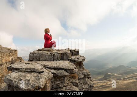 Ein wunderschönes Mädchen meditiert sich in einer lotus-pose, die vor dem Sonnenuntergang auf einem Felsen über den Wolken sitzt Stockfoto