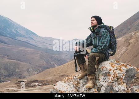 Müde bärtige Hüpfer mit Fernglas in den Händen sitzt auf einem Stein zwischen den Bergen und blickt in die Ferne Stockfoto