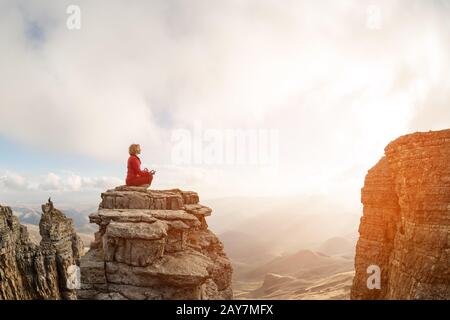 Ein wunderschönes Mädchen meditiert sich in einer lotus-pose, die vor dem Sonnenuntergang auf einem Felsen über den Wolken sitzt Stockfoto