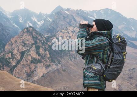 Ein Hüftkämpfer mit Bart in Hut, Jacke und Rucksack in den Bergen hält Ferngläser in den Händen und blickt in das Di Stockfoto