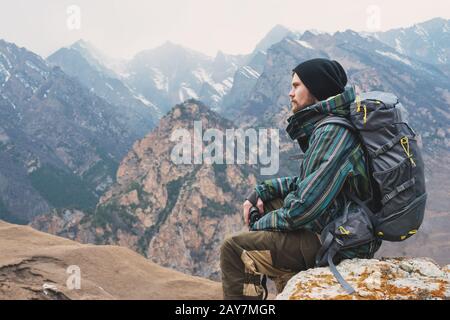 Müde bärtige Hüpfer mit Fernglas in den Händen sitzt auf einem Stein zwischen den Bergen und blickt in die Ferne Stockfoto