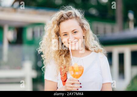 Horizontaler Schuss von hübsch geschweiften jungen Frauen mit charmantem Lächeln, hält Glas frischen Orangencocktail, schlendern im Freien, steht gegen verschwommenes Bac Stockfoto