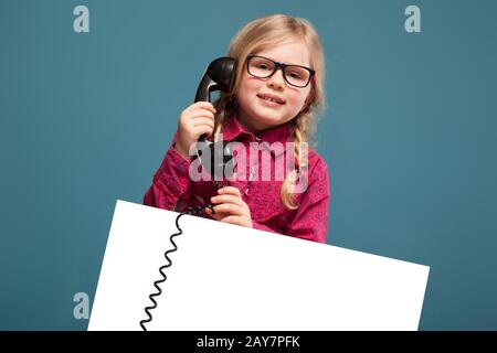 Hübsches, niedliches kleines Mädchen in rosafarbenem Hemd, schwarzer Hose und Brille hält leeres Plakat und hat ein Telefongespräch Stockfoto