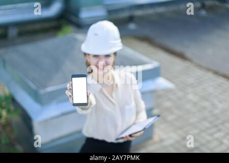 Attraktive Geschäftsfrau in weißer Bluse, Armbanduhr, Helm und schwarzem Rock stehen auf dem Dach und zeigen leeres Smartphone Stockfoto