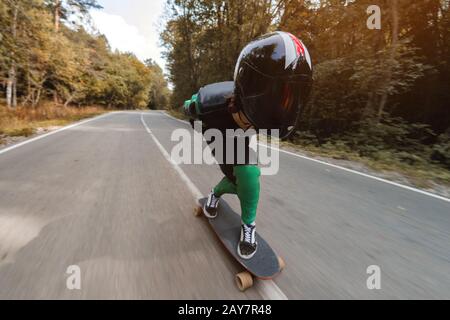 Ein junger Fahrer in einem speziellen Lederanzug und ein halber Helm fahren mit hoher Geschwindigkeit auf seinem Langbrett auf einer Landstraße im Wald Stockfoto