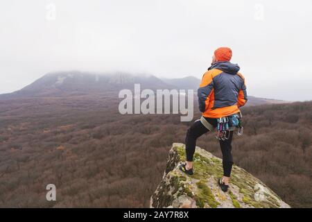 Hipster - ein Kletterer in einer Daunenjacke und einem gestrickten Mützenständer und ruht auf der Oberseite eines Felsens Stockfoto