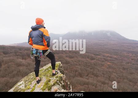 Hipster - ein Kletterer in einer Daunenjacke und einem gestrickten Mützenständer und ruht auf der Oberseite eines Felsens Stockfoto