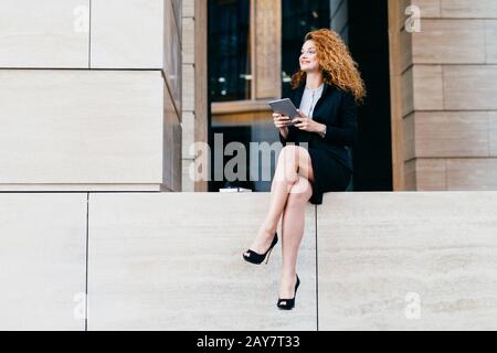 Porträt einer eleganten, prächtigen Frau in schwarzem Anzug und hoch gestielte Schuhe mit schlanken langen Beinen, mit modernem Tablet sitzend, mit fröhlichem Blick beiseite Stockfoto