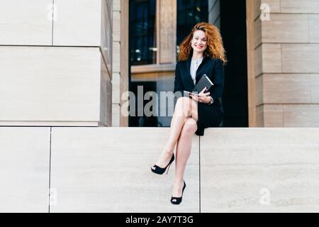 Junge attraktive Dame mit lockigen Haaren, mit schwarzem formalem Kostüm und Schuhen mit hohen Absätzen, mit Notizbuch und Tablet, die Arrangements Wi Stockfoto