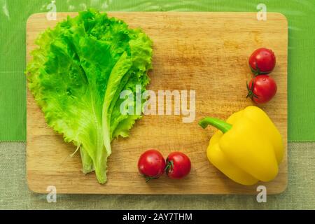 Frische grüne Salatblätter liegen auf einer Holzschneideplatte neben dem gelben Paprika und roten Kirschtomaten auf einem grünen Tisch Stockfoto