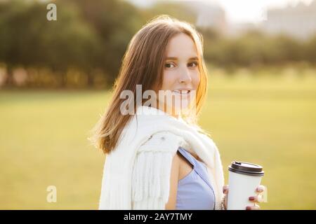 Porträt einer gut aussehenden jungen Frau mit ansprechendem Look, genießt aromatischen Kaffee aus Einwegbecher, schlendern mit Freund im Feld, sieht glücklicher aus Stockfoto