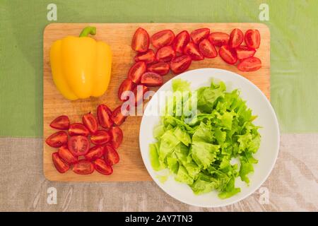 Frische gehackte grüne Salatblätter liegen auf einer Holzschneideplatte neben dem gelben Paprika und gehackten roten Kirschtomaten Stockfoto