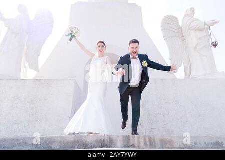 Portrait eines netten Paares von Neuvermählten an einem Hochzeitstag mit einem Blumenstrauß in den Händen, die die Treppe hinunterlaufen, lachend und lächelnd a Stockfoto