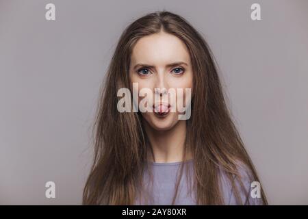 Lustige brünette Frau mit atraktivem Look, zeigt Zunge, macht Grimace und hat Spaß im Innenbereich, sieht mit blauen Augen auf Kamera, hat dunkle gerade Haare, iso Stockfoto