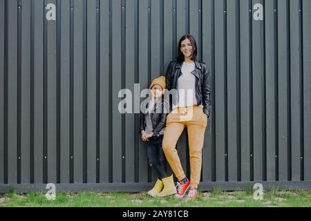 Modische, brünette Frauen in modischer Lederjacke und Sporthose, und ihre kleine Tochter steht in der Nähe, posieren an der Kamera gegen den Zaunbacken Stockfoto