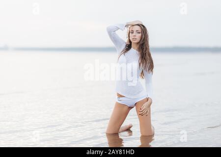 Outdooor Blick auf die gut aussehende brünette Frau mit Make-up, in weißen Bikini gekleidet, posiert bei der Kamera in der Nähe des ruhigen Meeresgrundes, hat eine passende Körperform, Gebühr Stockfoto