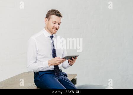 Positiver Finanzierer trägt weißes Hemd, Krawatte, sitzt auf dem Tisch, installiert neue Anwendung auf dem Touchpad, verbunden mit drahtlosem Internet. Erfolgreiche Busin Stockfoto