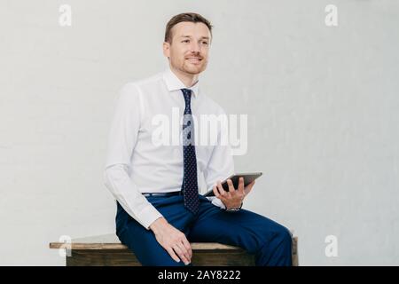 Nachdenklich attraktiver Geschäftsmann denkt über neue kreative Idee nach, verwendet modernes Touchpad, elegant gekleidet, sitzt allein auf Holztisch, posiert dagegen Stockfoto