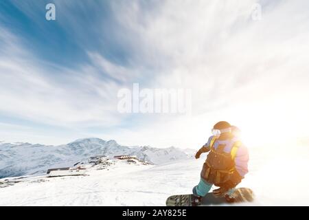 Ein Snowboarder in einer Skimaske und einem Rucksack fährt auf einer schneebedeckten Piste, die hinter einem Schneepulver gegen den blauen Himmel A zurückbleibt Stockfoto