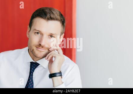 Selbstbewusster, unrasierter, männlicher Büroangestellter in formalem weißen Hemd mit Krawatte, hält Hand auf Wange, denkt darüber nach, eine neue Bühne im Leben zu beginnen, stellt sich gegen r. Stockfoto